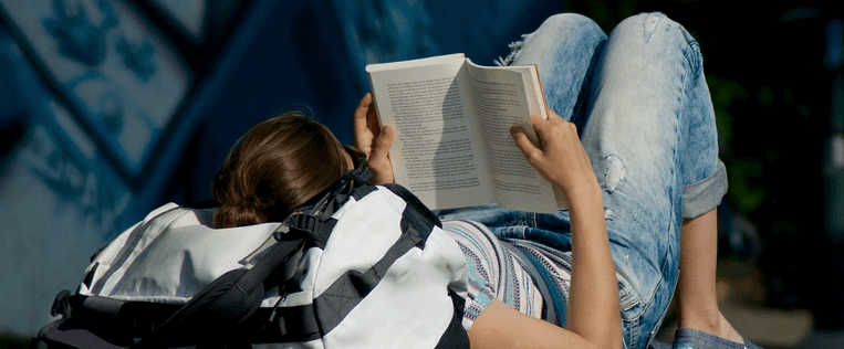 Pessoa lendo um livro deitada em cima de uma mochila 